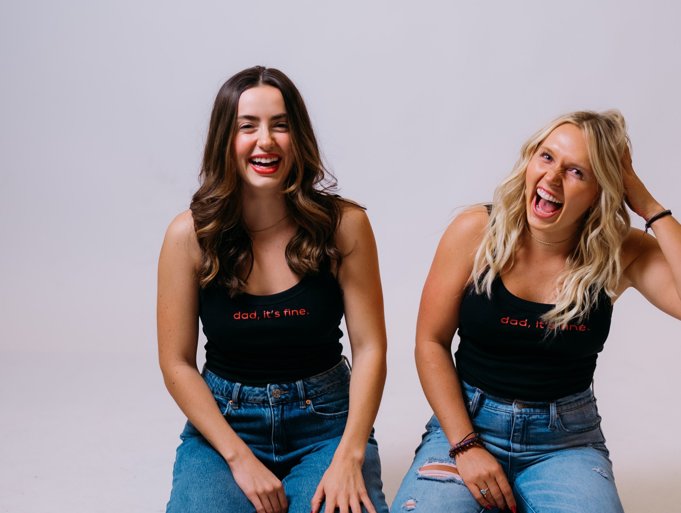 Two women sitting on the floor wearing matching black tank tops and blue jeans against a plain background. Dad its fine Austin Texas.
